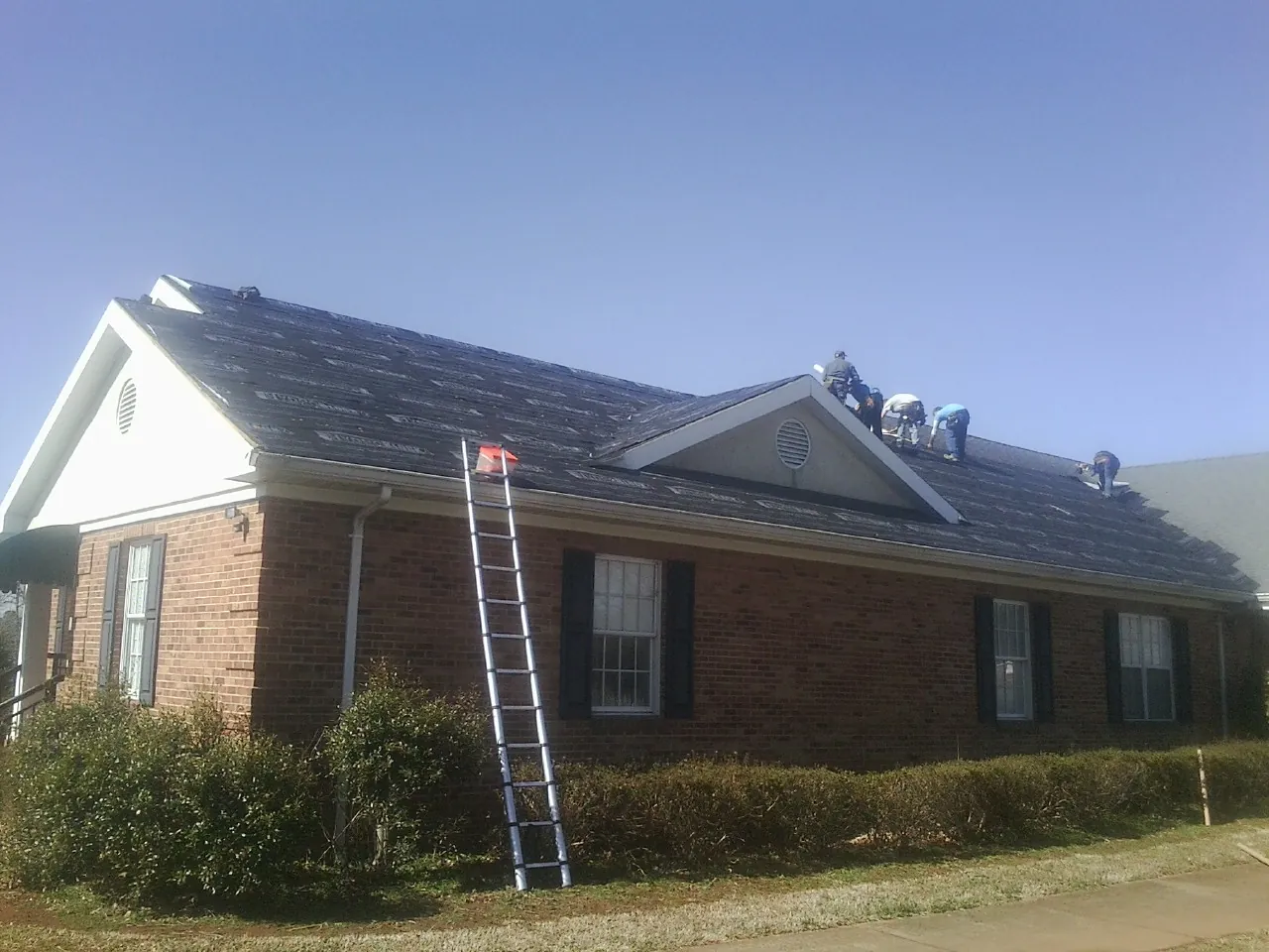 Crew tearing shingles off a brick ranch roof with a ladder set — Emergency Roof Repair work by our crew in Ambridge