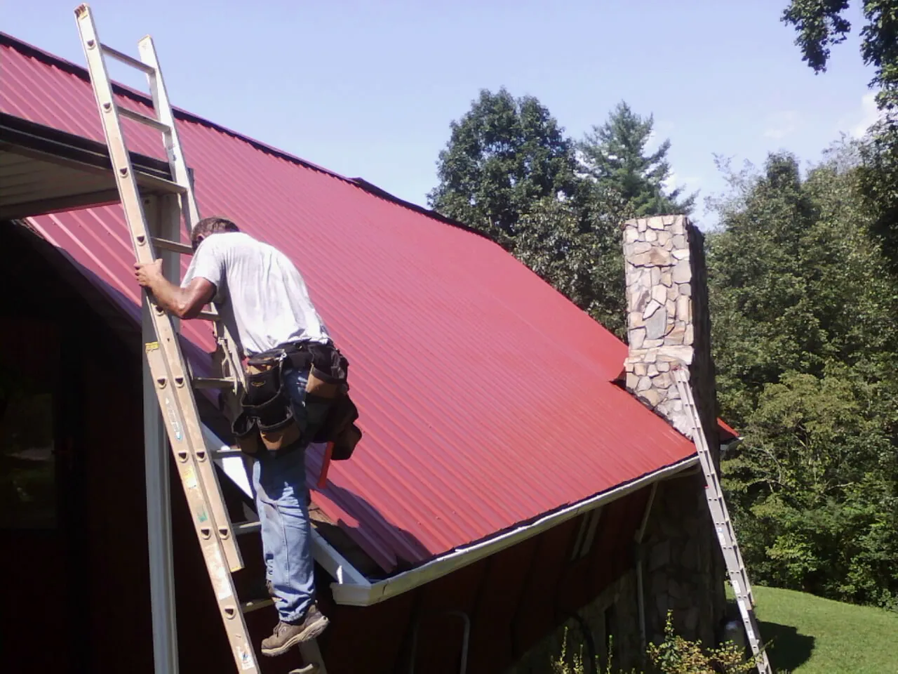 Roofer on a ladder beside a red metal roof and stone chimney — Soffit & Fascia Repair by Roofing Brothers in Ambridge